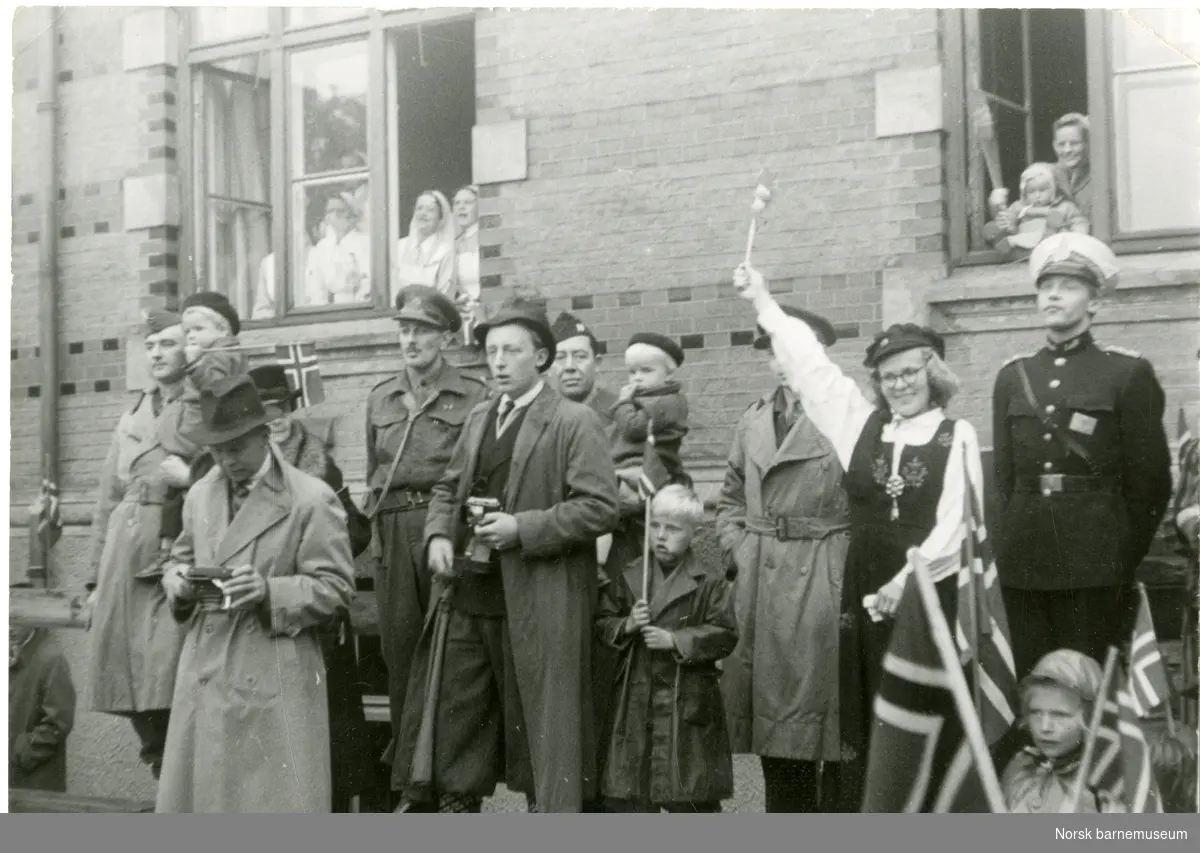 Festkledde mennesker med flagg utenfor Stavanger Sykehus 17. mai 1945. I vinduet til venstre er det flere sykepleiere. Jenta i vinduet til høyre er Astrid Lærdal (pikenavn) og hennes mor. Damen i bunad er hennes tante Hanna Marie Eskeland og mannen til høyre for henne i politiuniform er hennes onkel Odd Galtung Eskeland. To personer er i militær uniform. En person kikker på et kamera.