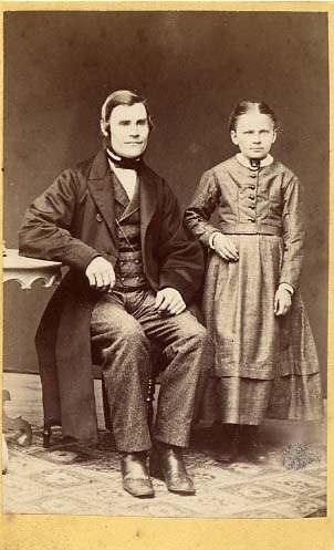 En man sitter på en stol med armen vilande på ett bord. En flicka står intill: "Hildur Holst med sin fader".