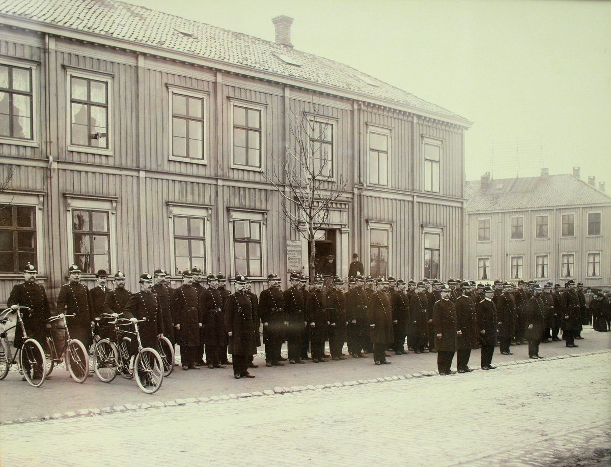 Trondheims politistyrke oppstilt foran politikammeret i Hornemannsgården på torvet i Trondheim. Alle er iført uniformsmodell 1895, og noen har sykkel.