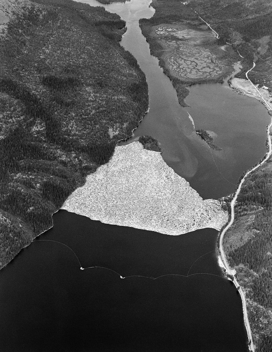 Flyfotografi fra nordenden av Sennsjøen i Trysil.  Bildet er tatt våren 1986 over et landskap med en smal innsjø omgitt av et terreng med skog, i hovedsak glissen bjørke- og furuskog, men også tettere granbestand i partier med djupere jordsmonn og mer fuktighet.   Langsmed østsida av vassdraget (til høyre i bildet) gikk Sennsjøvegen (fylkesveg 581) forbi Sennsjøvik-gardene.  Ute i innsjøene ser vi de to Sandholmene.  Like sør for disse holmene lå det ei lense, der tømmeret som var fløtet ned Femundselva fra Engerdal ble holdt tilbake i påvente av høvelig vannstand i den nedenforliggende delen av Trysilelva.  I den øvre delen av vassdraget var man avhengige av å fløte mens det var flomvannføring, for å slippe at tømmeret satte seg fast på grusører i elveløpet. Lengre nedover var det et poeng at vårflommen skulle være på retur før de store tømmermengdene kom. I det flate lendet langs Trysilelva kunne nemlig flomvannet føre mange av stokkene langt inn på land, og i så fall ble det arbeidsomt for fløterne å bære dem tilbake til vannstrømmen i elveløpet når vannstanden sank. Derfor ble engerdalstømmeret ofte holdte tilbake i Sennsjølensa til begynnelsen av juni. Det året dette fotografiet ble tatt ble det levert om lag 22 000 kubikkmeter fløtingsvirke fra Engerdal. Dermed ble bilvegene i regionen spart for 3 - 4 000 lastebillass. Fløterne var avhengige av nordavind når det åpnet Sennsjølensa, for strømdraget var ikke kraftig nok til å dra tømmeret gjennom sjøen om det blåste fra sør. På vannspeilet ser vi hvordan fløterne hadde sikret tømmerbeholdningen ved å legge ei sikringslense utenfor hovedlensa (i forgrunnen på bildet) og ei lense som hindret tømmeret i å drive inn i Sennsjøvika (til høyre). Da dette fotografiet ble tatt var det kraftig sønnavær, og vinden hadde blåst tømmeret litt tilbake fra stopplensa.