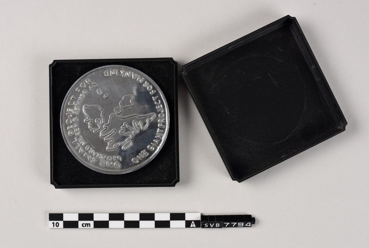 Skive av støpt metall med presset mønster og tekst på begge sider.
Medaljen ligger på en skumpute i en svart kunststoffeske med lokk .