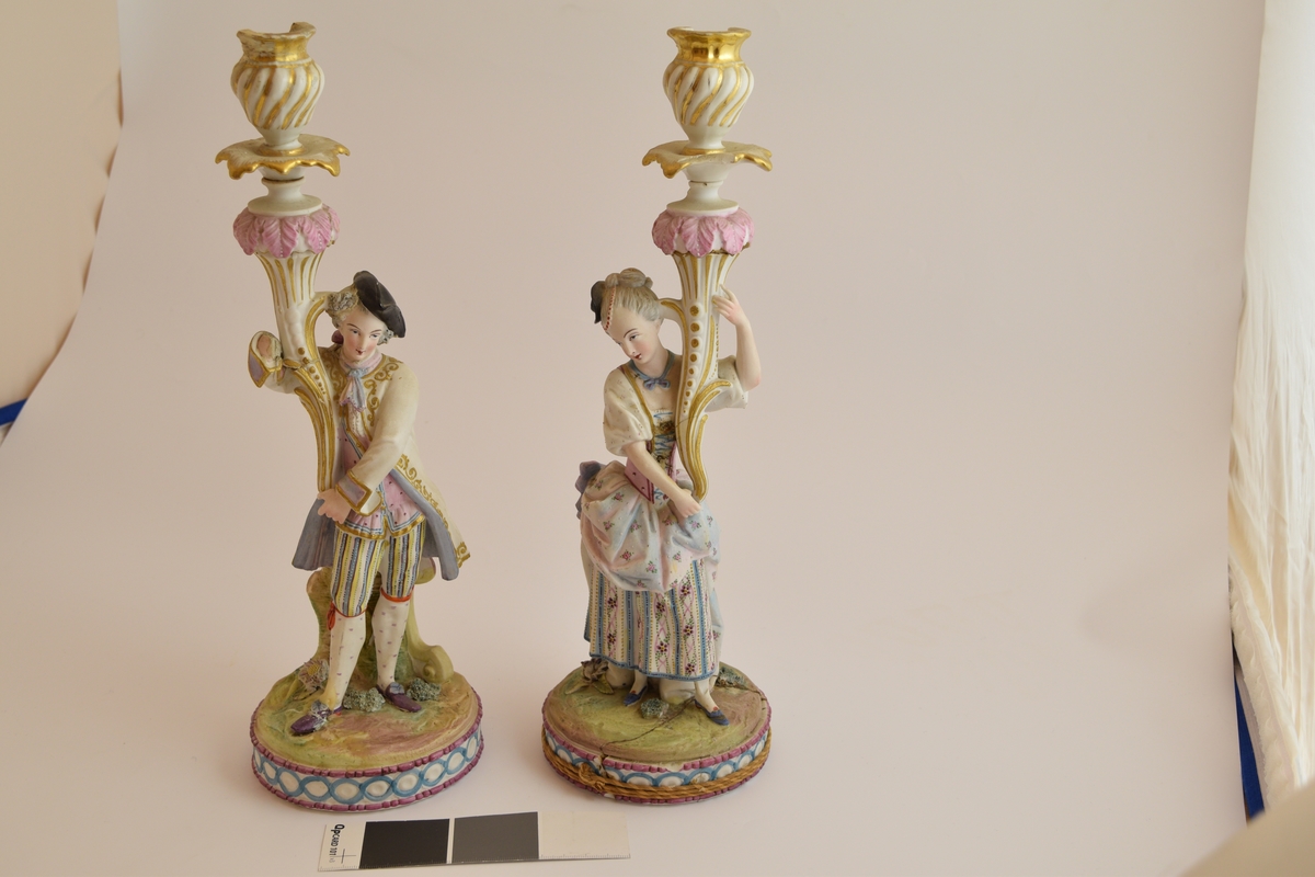 To lysestaker i form av statuetter av en mann og en kvinne i typisk 1700-talls fransk overklasse bekledning, De holder begge hver sin blomst som fungerer som stearinlysholdere.