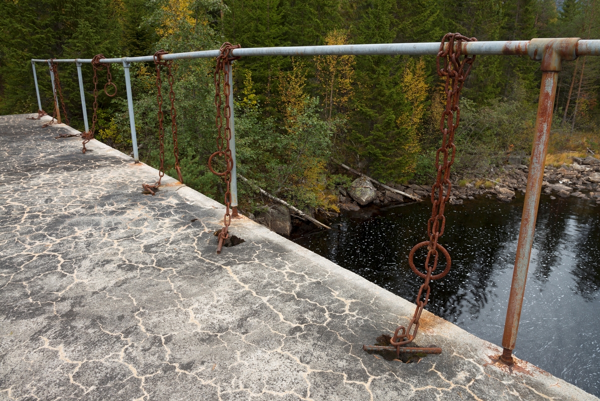 Detalj fra fløtingsdammen ved utløpet av Nedre Bjørvatn eller Nedre Bjorvatn [navneform brukt i dokumenter fram til midten av 1900-tallet] i Tinn i Øst-Telemark.  Den nedre Bjørvassdammen befinner seg høyt oppe i den fløtbare delen av Rauavassdraget.  Flere damkonstruksjoner har avløst hverandre på dette stedet.  Den siste ble støpt i 1962, som en såkalt «kvan-dam», med stålluker fra Foss bruk i Sande.  Fotografiet viser hvordan kjetting fra nederkanten av stållukene ble ført gjennom hull i den støpte brua, der det ble stukket jernteiner gjennom lekker i løftekjettingen når lukene sto i ønsket posisjon.  Løskjettingen ovenfor dette festepunktet ble knyttet fast til det galvaniserte jernrekkverket ved bruas ytterkant.  I bakgrunnen ser vi noe av vassdraget. 