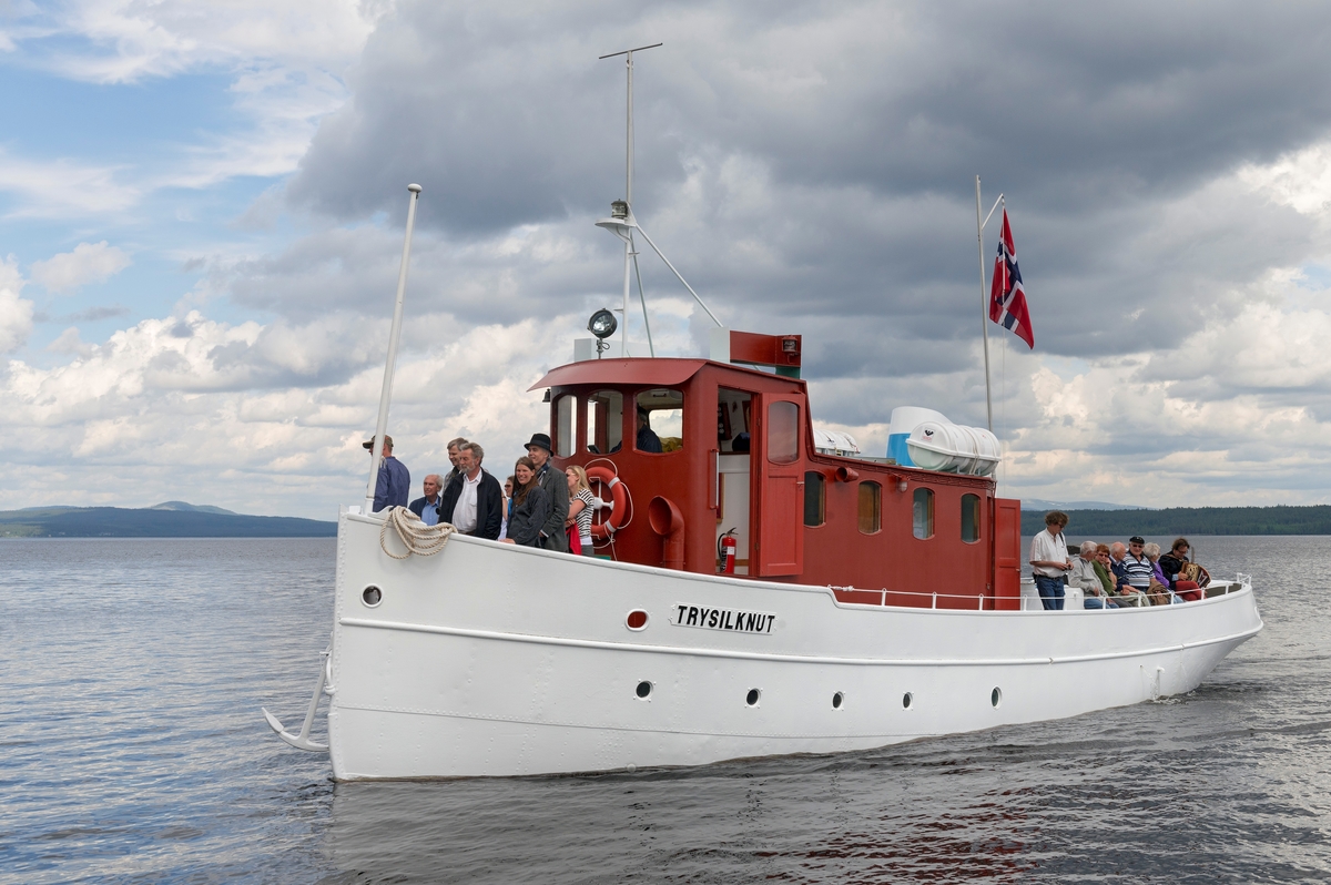 Slepebåten «Trysilknut» fotografert på den 47 kvadratkilometer store Osensjøen i grensetraktene mellom Åmot og Trysil i Hedmark, der Trysilknut gikk som tømmersleper fra 1914 til 1984.  Etter den tid har den 55 fot lange og 12 fot brede båten vært brukt som museumsbåt.  Den ble restaurert i perioden 2011-2014, og dette fotografiet er fra den første turen båten gjorde med passasjerer etter at restaureringa var avsluttet med maling av eksteriøret.  Skroget er kvitmalt og overbygningen (styrhus og bysse) er rødbrun.  Disse fargene har Trysilknut hatt siden båten ble ombygd fra damp- til dieseldrift i 1957-58, bortsett fra at vulsten som markerer hvor rekka møter den nedre delen av skroget lenge var grønnmalt.  Fotografiet er tatt i forbindelse med markeringa av båtens 100-årsjubileum 11. juni 2014.  På fordekket sto maskinist og omviser Allan Tutvedt [delvis skjult bak flaggstanga], pensjonert museumsdirektør Tore Fossum, konservator Bjørn Bækkelund fra Norsk Skogmuseum, direktør Harald Jacobsen fra Anno museum AS, fagkonsulent Ingvild Herberg og avdelingsdirektør Stig Hoseth, begge fra Norsk Skogmuseum.  Mer om Trysilknut og dens historie under fanen «Andre opplysninger».