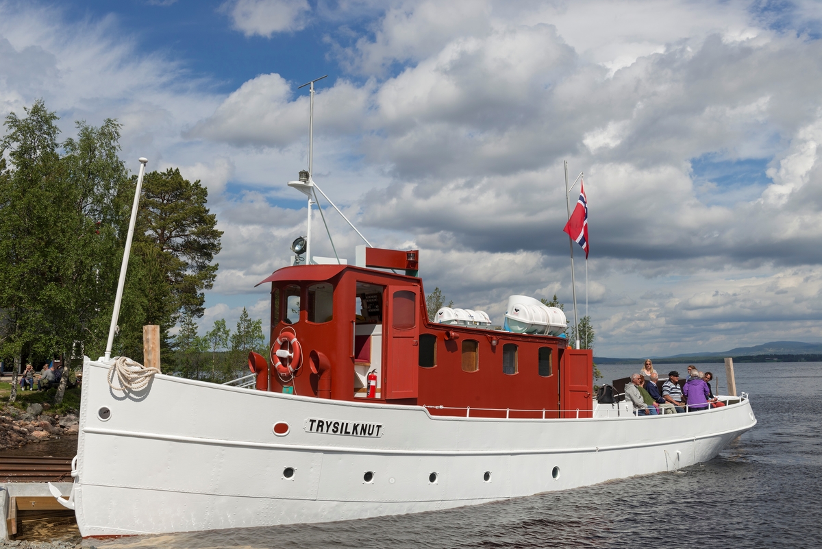 Slepebåten «Trysilknut» fotografert på den 47 kvadratkilometer store Osensjøen i grensetraktene mellom Åmot og Trysil i Hedmark, der Trysilknut gikk som tømmersleper fra 1914 til 1984.  Etter den tid har den 55 fot lange og 12 fot brede båten vært brukt som museumsbåt.  Den ble restaurert i perioden 2011-2014, og dette fotografiet er fra den første turen båten gjorde med passasjerer etter at restaureringa var avsluttet med maling av eksteriøret.  Skroget er kvitmalt og overbygningen (styrhus og bysse) er rødbrun.  Disse fargene har Trysilknut hatt siden båten ble ombygd fra damp- til dieseldrift i 1957-58, bortsett fra at vulsten som markerer hvor rekka møter den nedre delen av skroget lenge var grønnmalt og at Glomma Fellesfløtingsforenings initialer på skorsteinen skal ha hatt en gulere fargetone.  Fotografiet er tatt ved brygga på Sørlistøa fløtermuseum den 11. juni 2014, da museet markerte båtens 100-årsjubileum.  Mer om Trysilknut og dens historie under fanen «Andre opplysninger».