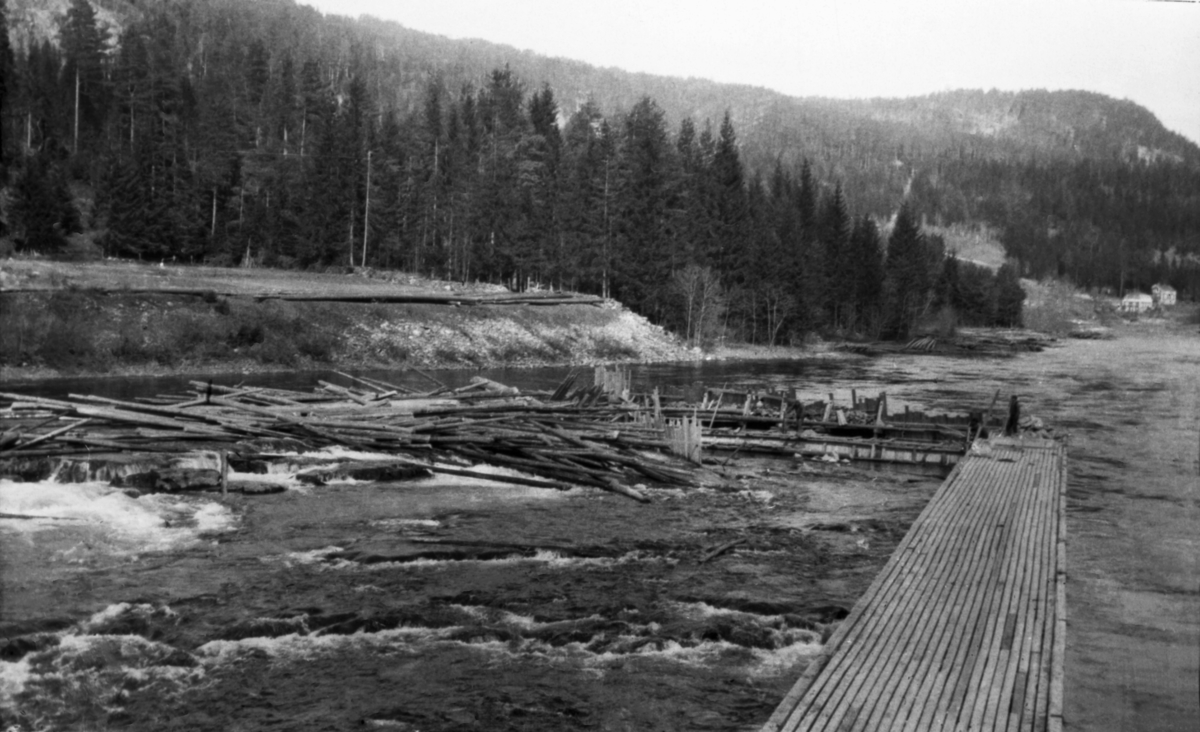 Tømmerhaug i Sagafoss i Tinnåa (Tinne) i Notodden (tidligere Heddal eller «Hiterdal») kommune i Telemark.  Dette fotografiet fra 1921 er tatt i motstrøms retning i den øvre delen av fossen.  Tømmeret har lagt seg på en avsats i den øvre delen av fossen, ved siden av en skjerm som er bygd sentralt i elveløpet, vinkelrett på strømretningen.  Opp til denne skjermen går det ei gangbru av tre.  Høyere oppe ved vestsida av elveløpet skimter vi mer fløtingstømmer langs elvebredden.  Sagafossen var om lag ti meter høy, og den voldte atskillige ulemper for fløtinga i Tinnåa, for det hadde lett for å sette seg tømmerhauger under dette vannfallet, både når det var høy og når det var lav vannføring.  I slutten av 1890-åra ble det bygd tømmerskjermer på begge sider av fossen, noe som skal ha redusert problemet.  Disse skjermene er imidlertid ikke å se på dette fotografiet.  Sagafossen ble helt borte da den nye Tinnfossdammen ble tatt i bruk i midten av 1950-åra. 