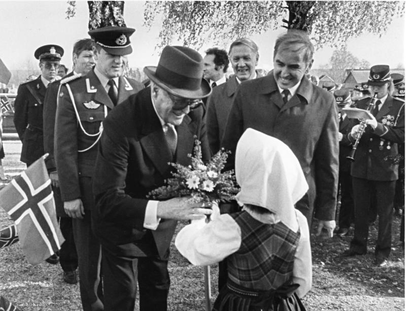 En mann med hatt mottar blomster av en ung jente. Omkring dem er mennesker i penklær og norskeflagg.
