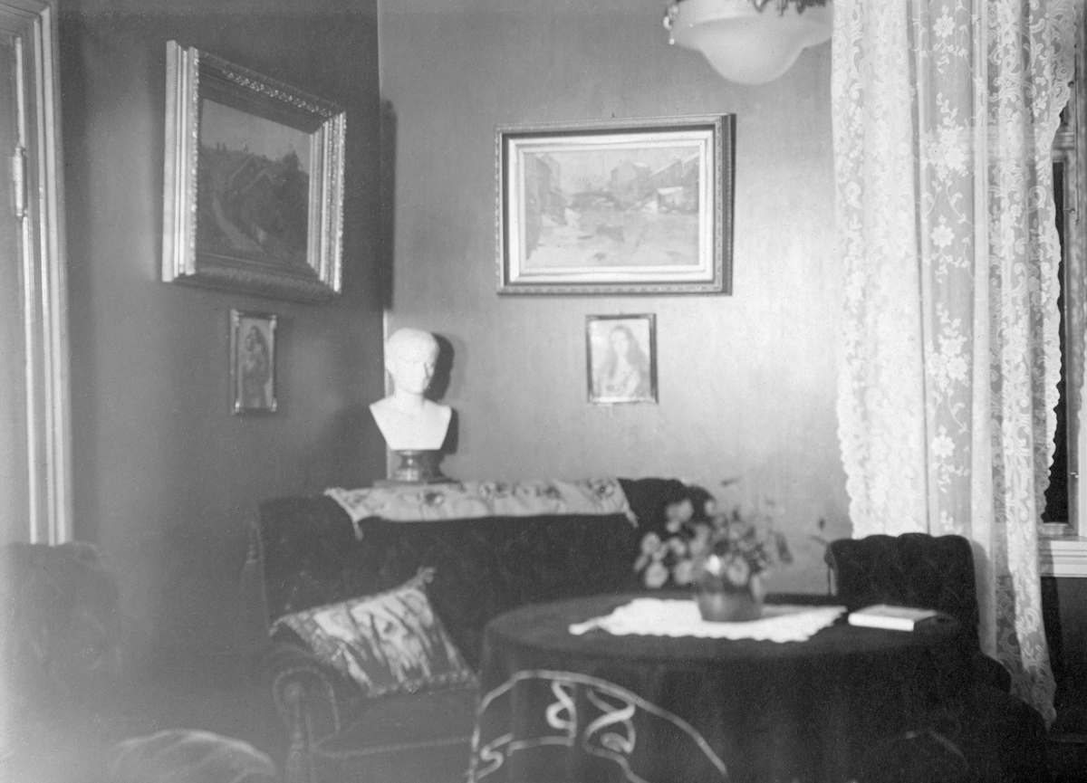 Interiør fra stua i lensebestyrerboligen på eiendommen Lund ved Fetsund.  Bildet viser et forholdsvis lyst rom med tapetserte vegger.  Til høyre ser vi et stort krysspostvindu med kniplingsgardiner, til venstre en dørkarm mot et tilstøtende rom.  Bildet viser ellers et hjørne med plysjsofa og to lenestoler mot et rundt bord med en mørk duk på.  Oppå duken, sentralt på bordflata lå det en mindre, kvit duk, som det sto ei blomsterpotte på.   I hjørnet bak sofaen sto det en skulptur, et kvitt hode, antakelig en gipsetterlikning etter et antikt forbilde. Veggene var prydet med to malerier og to mindre bilder.  Bildet skal være fra 1919. Dette året flyttet fløtingsdirektør Johs. Johannesen, kona Harriet og barna deres fra Fetsund til Vindern i Kristiania. Etter dette var det lensebestyrerne Martin Kveseth og Alfred Bjølgerud samt fullmektig Gabriel Faye og familiene deres som bodde i våningshusa på Tømmerdirektionens eiendom Lund.