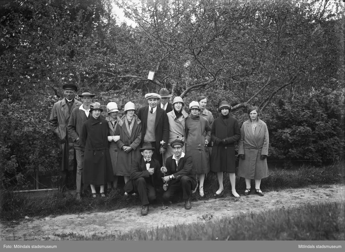 Tretton kvinnor och män står uppställda vid en smal grusväg och två män sitter på huk framför. Samtliga är snyggt klädda och några använder hatt. I bakgrunden ser man träd och buskar. Troligtvis 1920-30-tal.