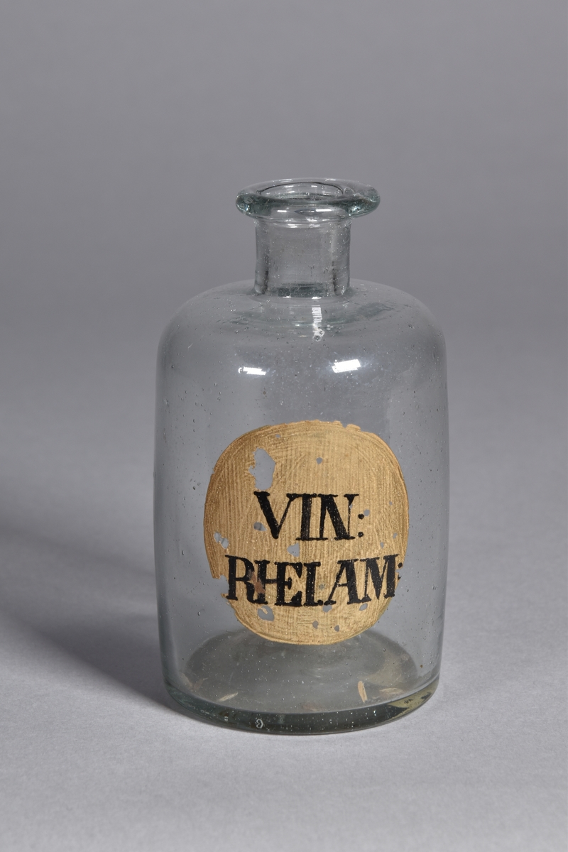 Flaska av klart glas, cylindrisk, kort hals med utsvängd mynning. Påmålad etikett med svart text.