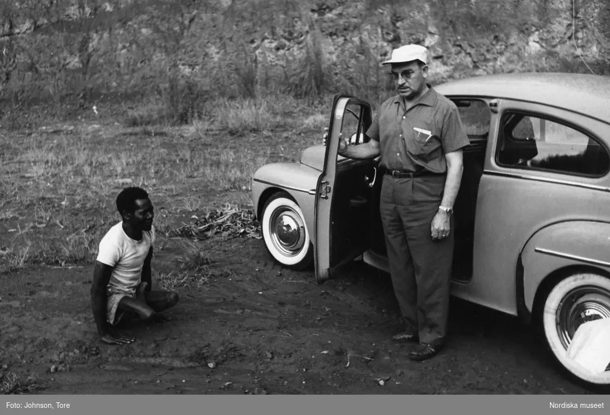 Kongo. En FN-tjänsteman står vid sin bil, en Volvo PV 544. Bredvid honom en sittande kongolesisk man.