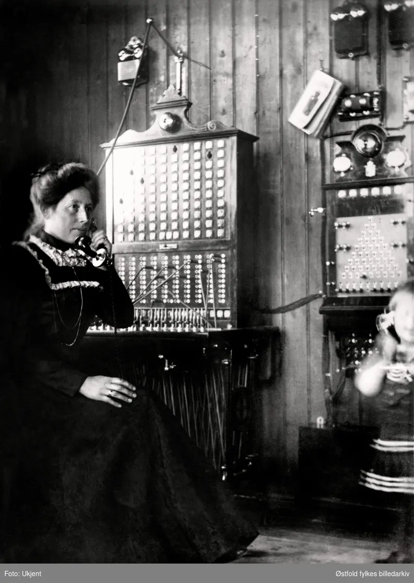 Rakkestad Telefonsentral 1902. Til venstre telefonistinde Mina Torper ved sentralbordet. Sønnen Christoffer, tre år er med mor på jobb.