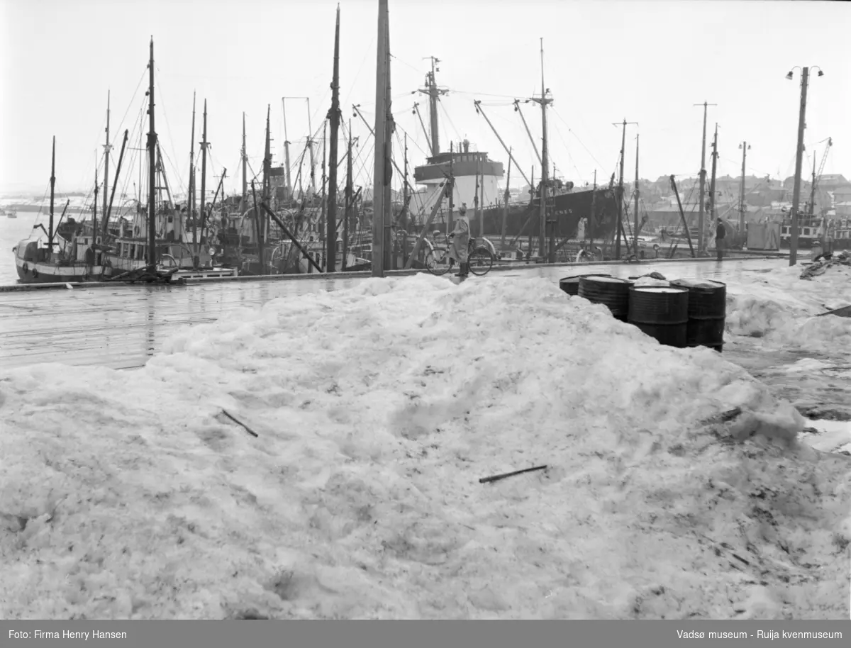 Vadsø havn 1952. Bildet er tatt på kaia mot nordvest, delvis snødekt kai i forgrunnen. Fiskebåter, en mann med sykkel, to større fartøy ved dampskipskaias utstikker.