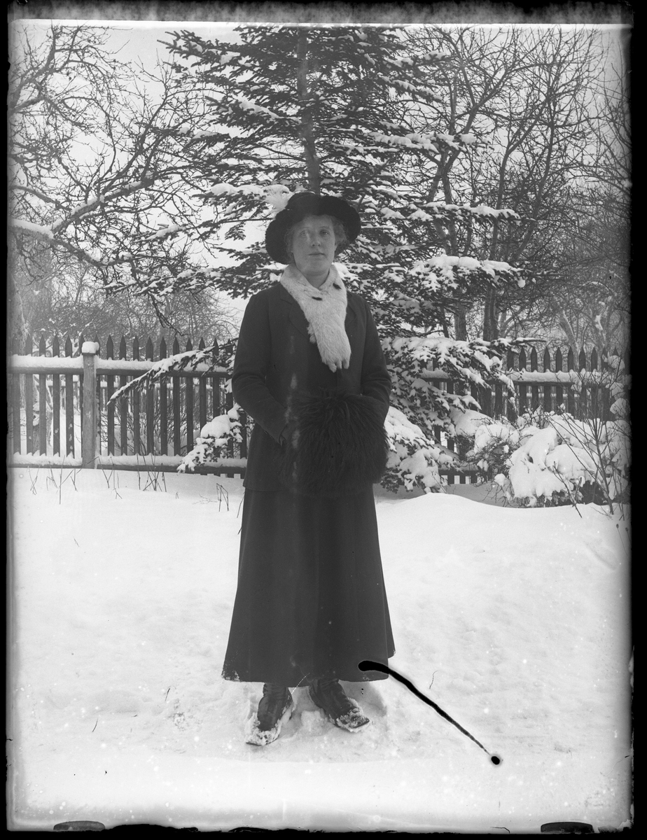 Hildur Wilhelmsson porträtterad i snön klädd i mörk kappa, svar pälsmuff, vit pälskrage och svart bredbrättad hatt med en vit fjäder.