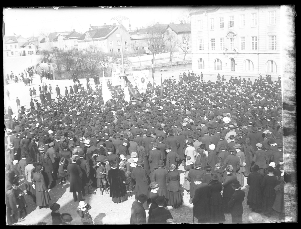Första maj-demontration på Lilla torget. Kvinnor, män och barn i mörka kläder har samlats på torget och står vända mot Stora torget.