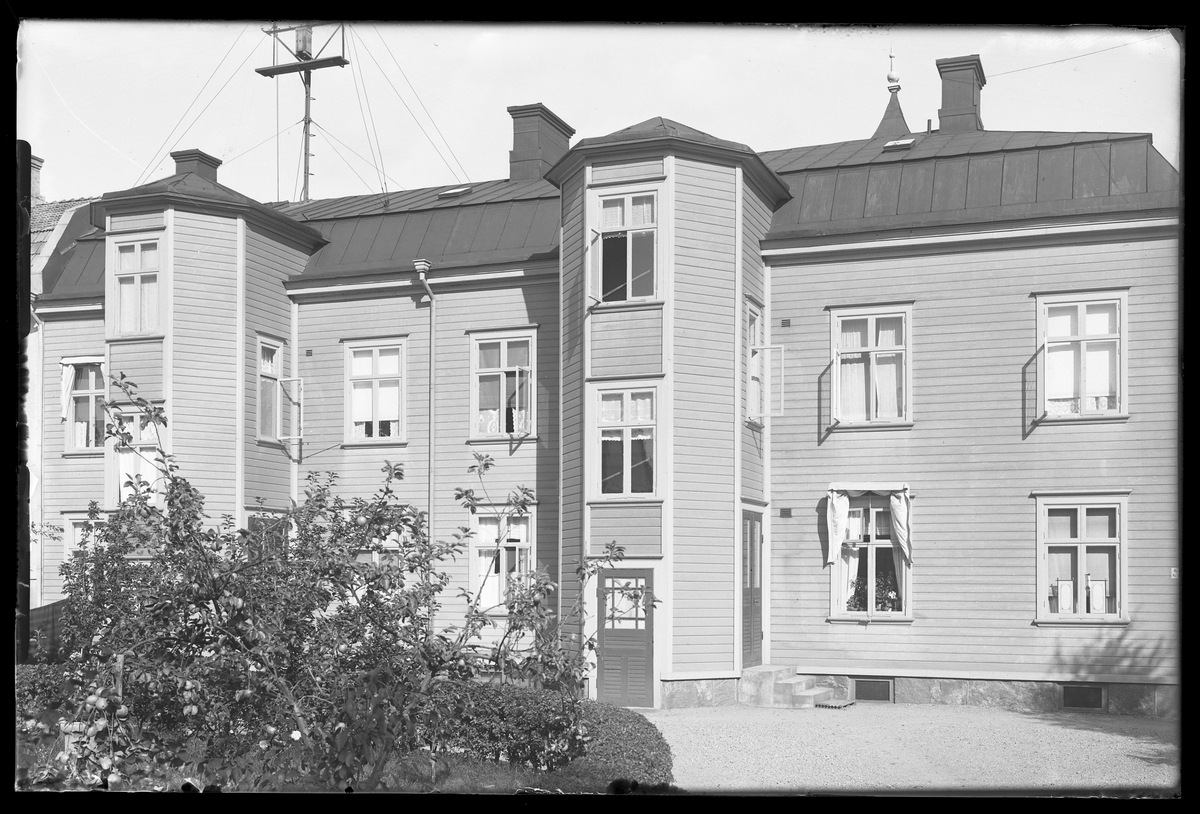 Ett lägenhetshus med liggande panel och plåttak. På huskroppen finns två utstickande trapphus och framför ligger en grusgång och buskar. I fotografens anteckningar står det "Fam Wincrant hus".