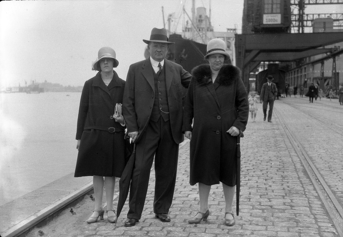 Två kvinnor och en man står vid vattnet i en hamn, troligen Frihamnen i Göteborg. I bakgrunden syns människor, ett fartyg samt byggnader. I fotografens egna anteckningar står det "Maggnussons, Gbg".