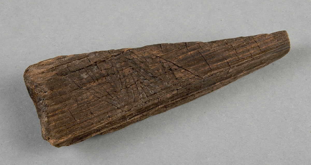 Bearbetat trä. Triangulärt föremål med rektangulärt tvärsnitt, trolig möbeldetalj. Dekorerad med linjeornamentik.