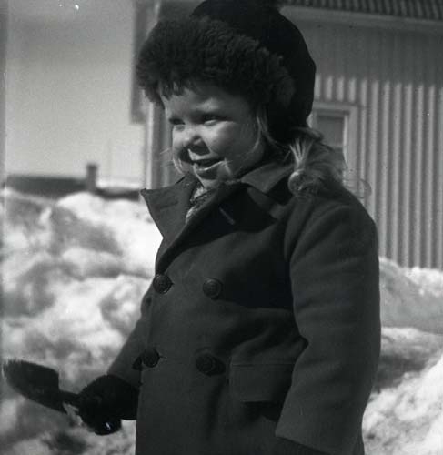 En flicka står utomhus klädd i kappa och pälsmössa. I ena handen har hon en spade, 1947.