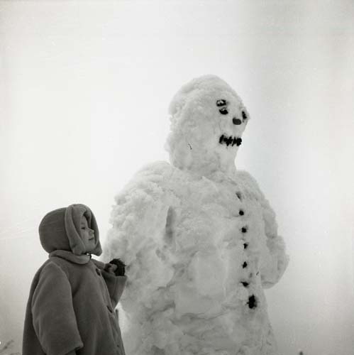 En flicka iförd en tjock kappa står intill en leende snögubbe, 1955.