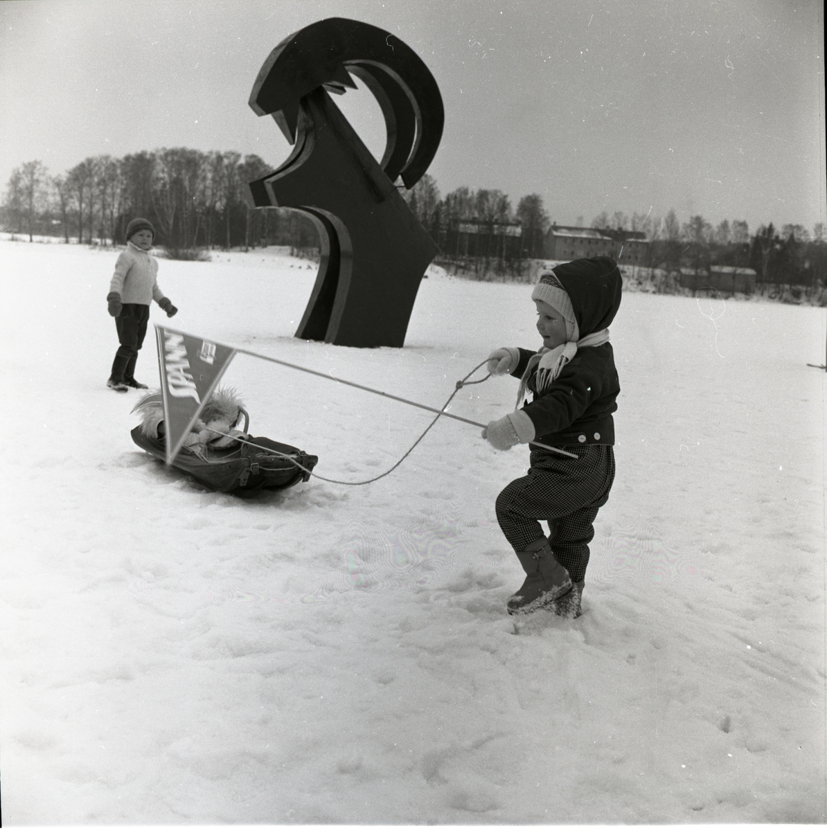 Ett barn drar en kälke förbi en hälsingebock vid svenska mästerskapen i budkavle den 12-13 mars 1960 i Bollnäs. Intill bocken står ytterligare ett barn.