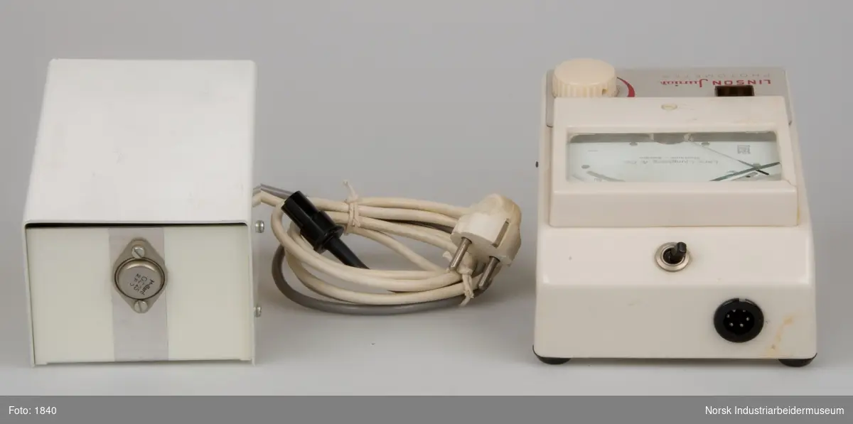 Måleinstrument med analog viser og tilhørende transformator. Instrumentet er et fotometer for hemoglobinundersøgelser og kan måle mængden af lys der trænger gennem en væske. Transformatoren er en 220 volt enhed og er forsynet med en ledning isoleret med blød plast.