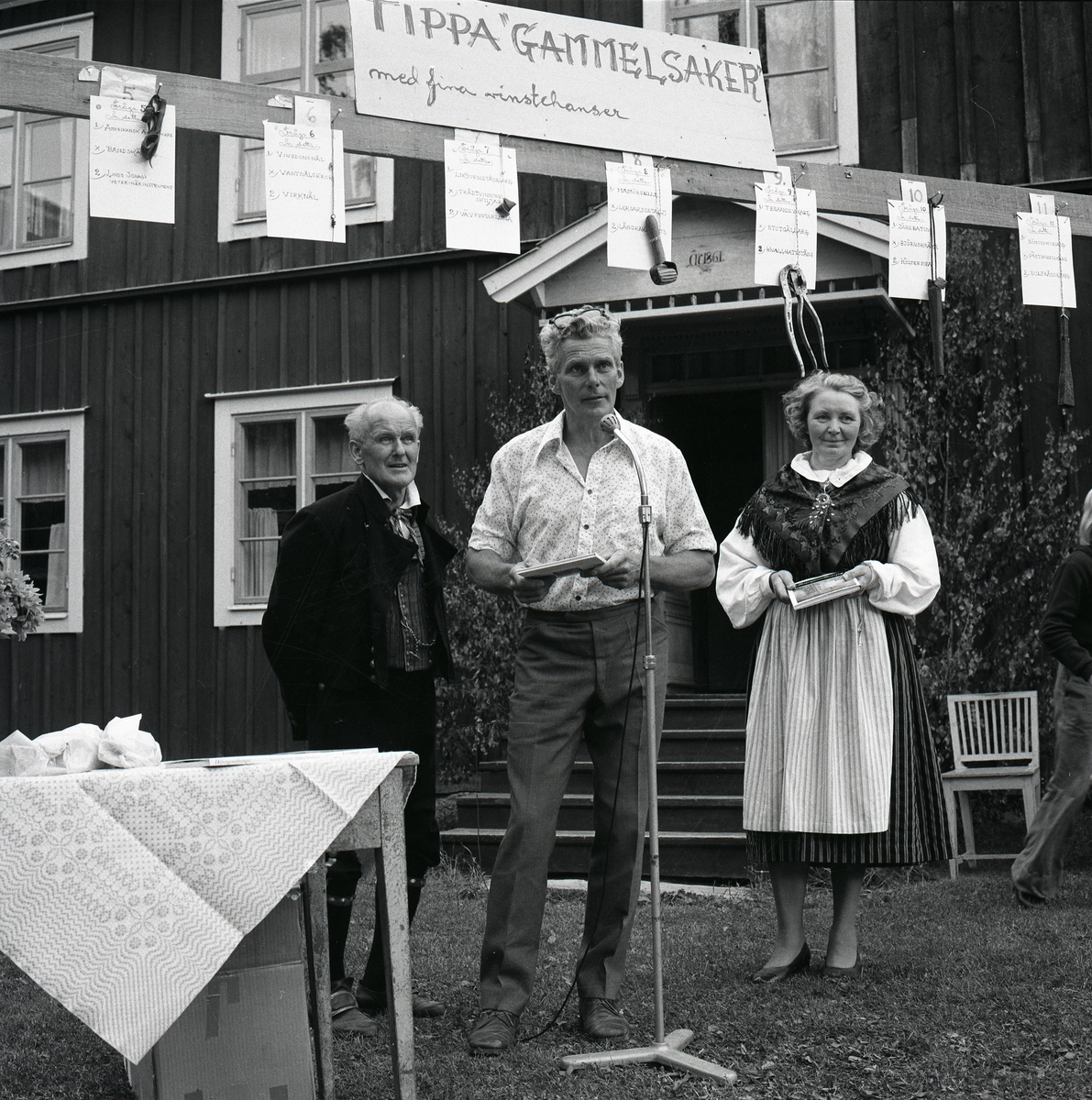 Två män och en kvinna står vid en mikrofon under julifesten vid Hembygdsbyn, 1975. Ovanför deras huvuden hänger föremål som ingår i en tävling.
