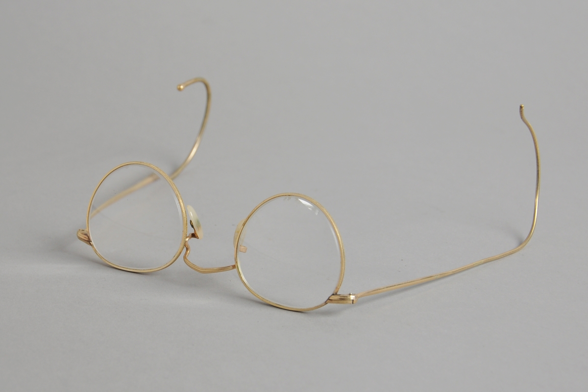 Brillen har tilnærmet sirkelrunde glass med sterk pluss-styrke, og gullfarget metallinnfatning og stenger som er buede ytterst.