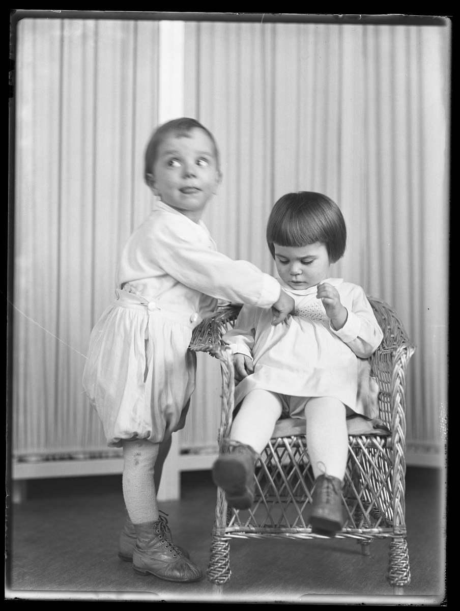 En flicka sitter i en korgstol medan hennes bror står bredvid. I fotografens anteckningar står det "Ing.[enjör] Bergs barn", tolkat som tvillingarna Peter Wilhelm och Ulla Maria.