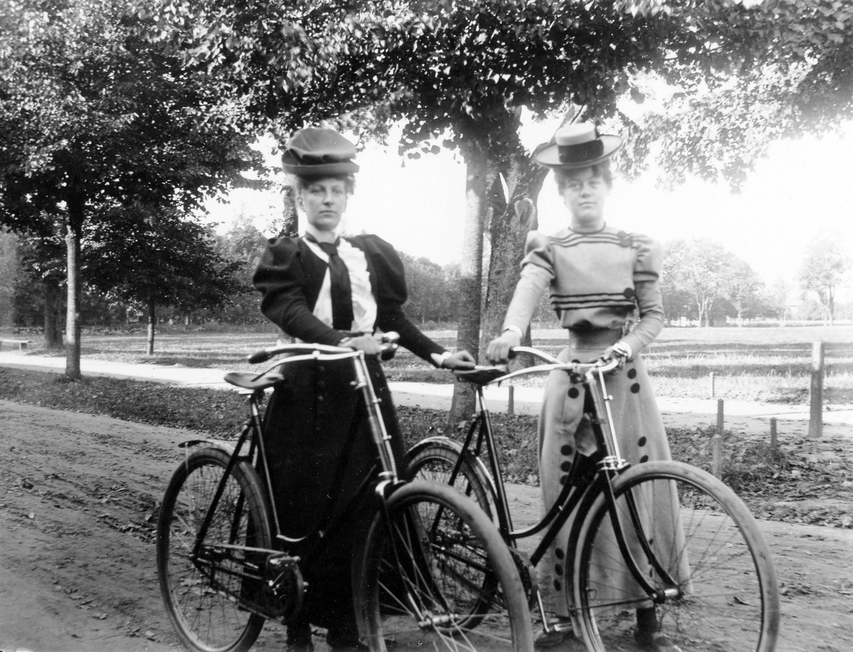 Två kvinnor på cykeltur, iklädda klänning och hatt, poserar bredvid varandra med var sin cykel på Norra Ringgatan.