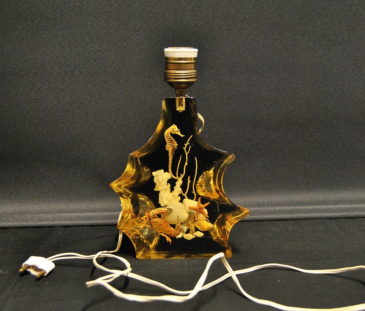 Lampe med uregulær form. Støpt i lucite recin akryl. Støpt i plasen er korall, sjøhest, krabbe og skjell. Er mest sannsynlig fra 1960-1980- årene. Mulig souvenir fra Spania.