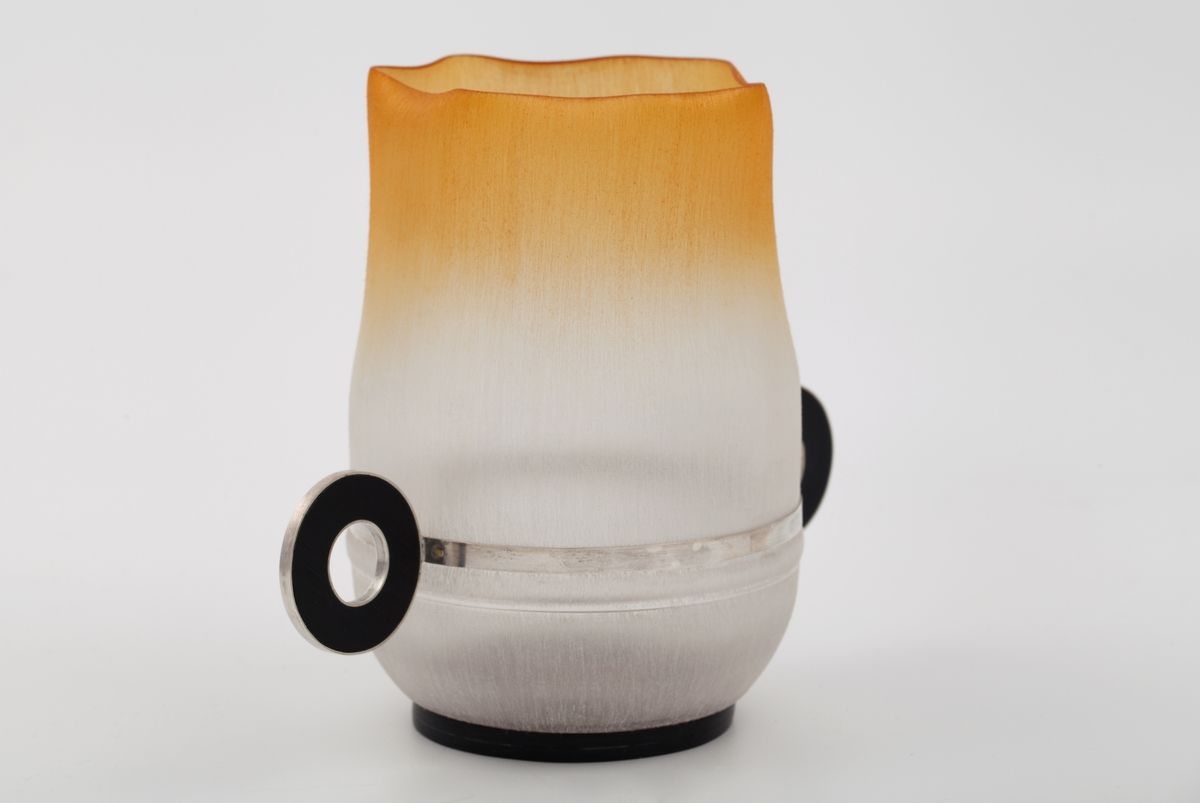 Objekt fremstilt av nedre del av en mineralvannsflaske, pusset med smergelpapir (pussepapir) og farget oransje. Bunnen og de små runde hankene på hver side er laget i sort hardplast og sølv.