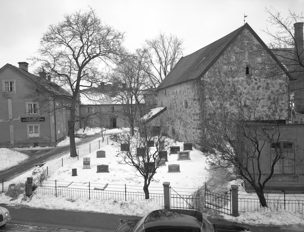 Vy mot Rhyzeliusgården i Linköping. Byggnaden till vänster uppfördes 1723-24 som domprostgård. Stenhuset till höger kopplades tidigare till en eventuell klosteranläggning på platsen. Den tolkningen har dock falsifierats i senare tid. Numera inrymmer byggnaden ett stenmuseum. Begravningsbyrån och gravstenarna till trots har tomten aldrig varit föremål för begravningar. Gravvårdarna förklaras av att Nelsons stenhuggeri under lång drev verksamhet från gården. Bild från omkring 1955.