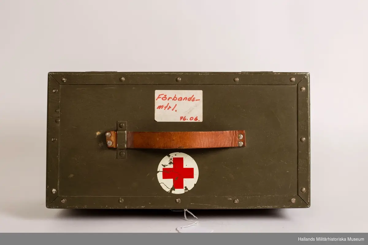 En låda i trä (41 x 16, 5 cm) med läderhandtag. Gröngrå färg med röda korsets emblem på. Lådan innehåller förbandspaket och diverse sjukvårdsmateriel. Vit klisterlapp ovanpå locket med röd text, handskrivet.
