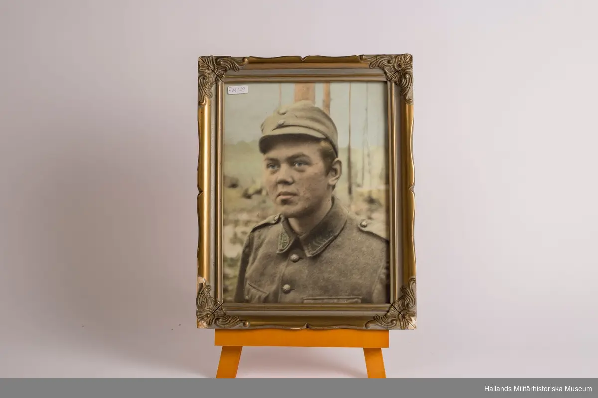 Fotografi i ram. Föreställande ett porträtt av soldat. Halvbyst. Dekorativt utsmyckad ram av trä, guldmålad.