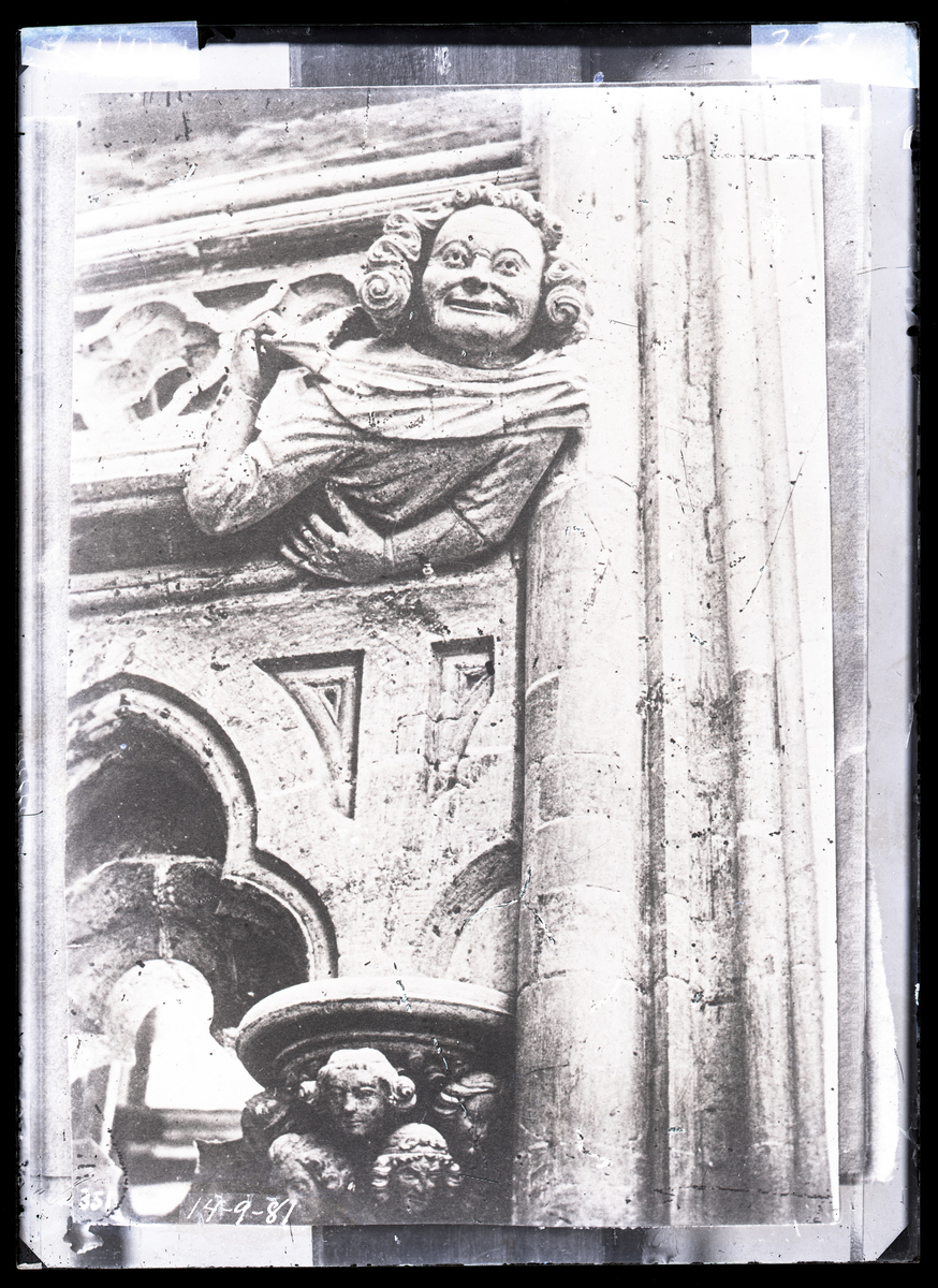 Hilsende figur i nord-vestre del av ambulatoriet i oktogonen i Nidarosdomen. Skulptur laget etter brannen i 1328.