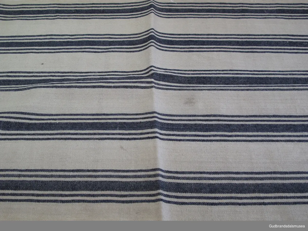 1 lengde vevd klede med stribet mønster, for dynetrekk til underdyne, dvs. madrass.. 1 av 3 kleder med likt stoff, veving og mønster med brede, vertikale striper i blått.
