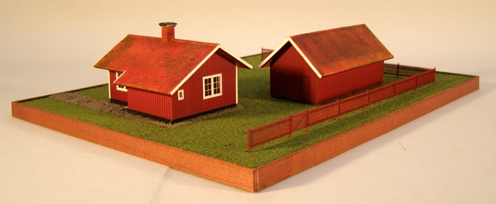 Modell av Banvaktstuga 213 Hosäter med uthus och staket,  i skala 1:87 H0. Husen är byggda av etsark i mässing och målade med modellfärg av märket "Model master". Basplatta av Mdf med strömaterial.