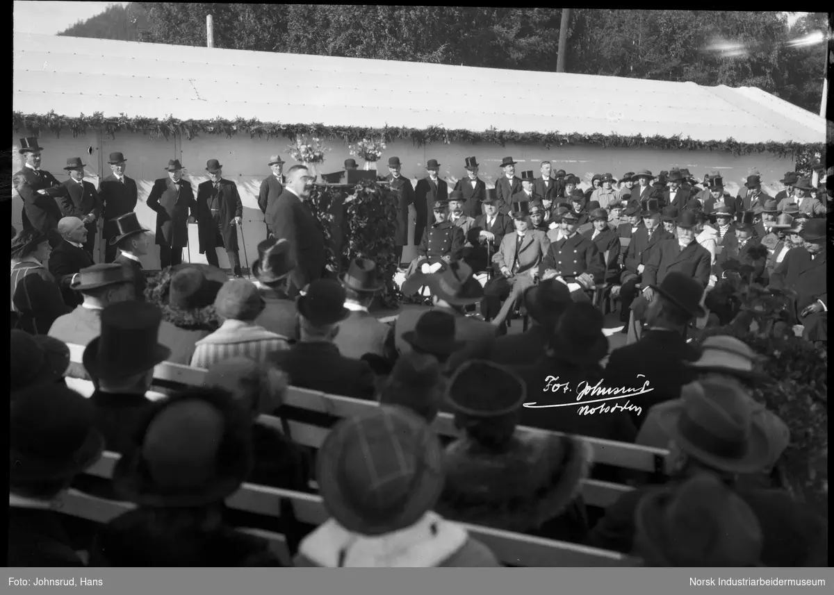 Åpning av Fylkesutstillingen 1922 med besøk av Kong Haakon VII. Person taler foran folkemengde. HM Kongen sitter som tilhører.