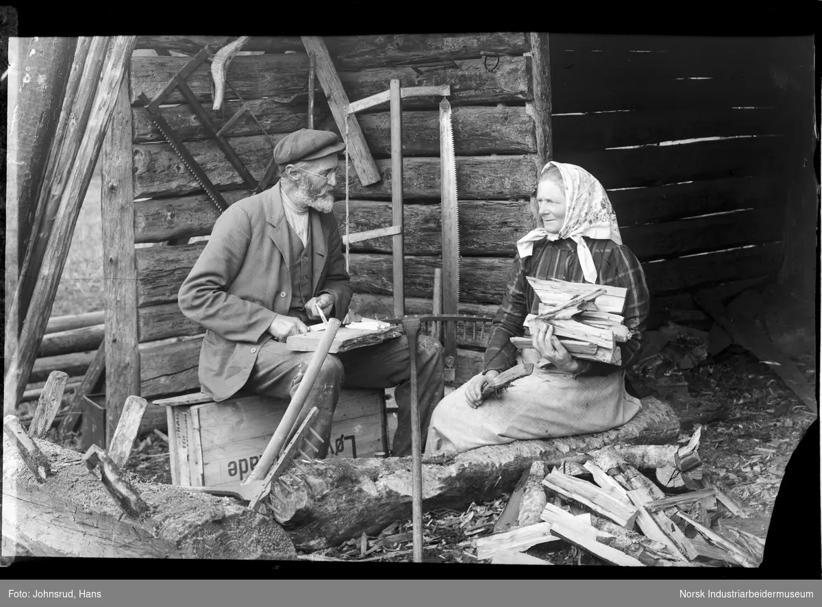 Ole og Berthe Marie Johnsrud sittende utenfor laftet vedskjul. Kvinnen med ved i armene, mann i arbeid med treskjæring.