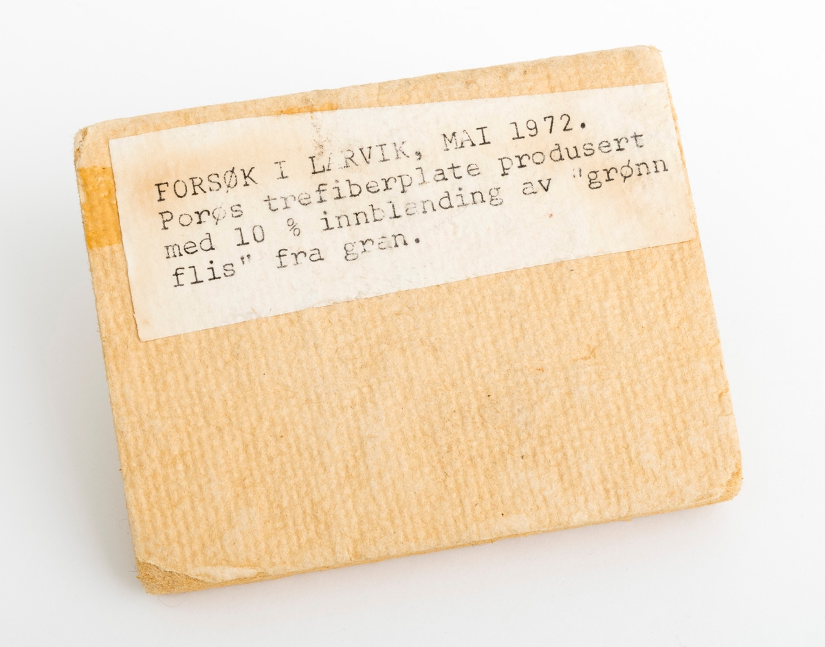 En rektangulær trefiberplate (wallboardplate), vareprøve, på 9,0 cm x 7,0 cm. Plata er porøs med et pålimt skikt av et tapetlignende materiale på framsida. På vareprøvas ene side gjenifnnes en påklistrert etikett med denne teksten: Forsøk i Larvik, mai 1972. Porøs trefiberplate produsert med 10 % innblanding av "grønn flis" fra gran.