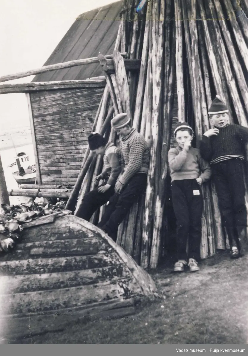 Fire gutter står lent mot råvedstabel foran båthus i Skallelv. Bl.a. Olaf Harjo, Edmund Pleym, og Are Harjo. Ca begynnelsen 1960-tallet.