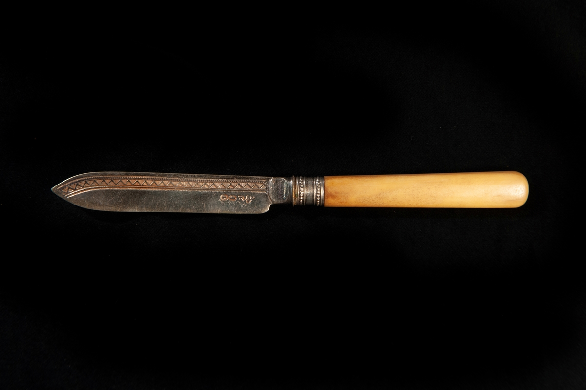 En fruktkniv av silver/nysilver med skaft i gulbeige ben. Dekor i form av zickzackbård på bladet. Otydliga utländska stämplar. Jfr JM 18876 - 18878.
