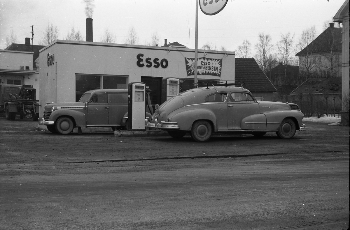 Ekspedisjonsbygg og bensinpumper på Esso, Lena, tidlig vår 1955. Bilene ved pumpene er iflg. informant en Opel Olympia varebil, årsmodell 1950-53, og en Pontiac (kjennetegn bred kromlist over bagasjelokket).