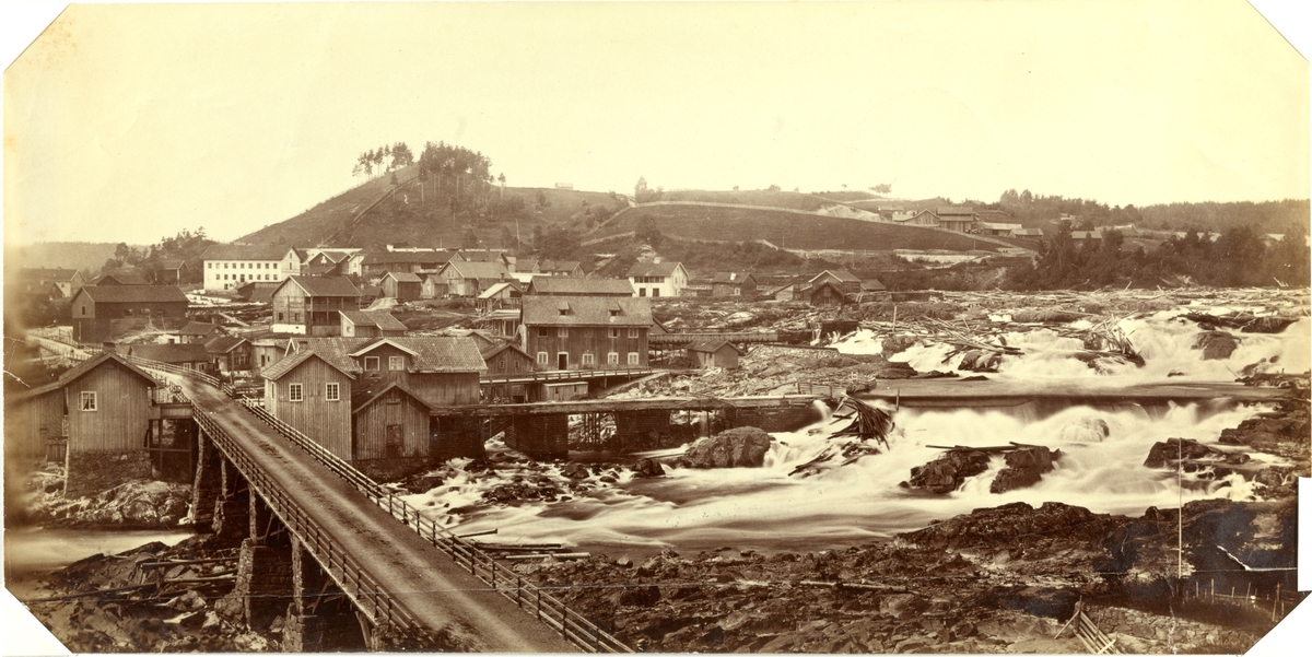 Hønefoss under tømmerfløtingen. Byen i bakgrunnen og bro over fossen i forgrunnen. Fotografert mellom 1870 og 1886.