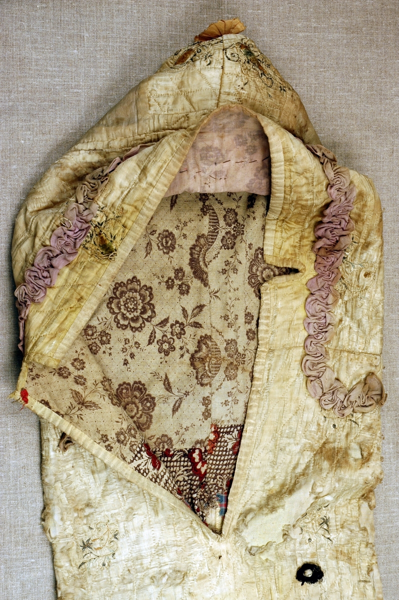 Dåpspose i brodert og vattert silke med stikninger, dekorert med blonde- og stoffrosetter med pynteknapper, perleblonde, rysjer og band. Foret med tre forskjellige stoffer, med ruter og blomster.
