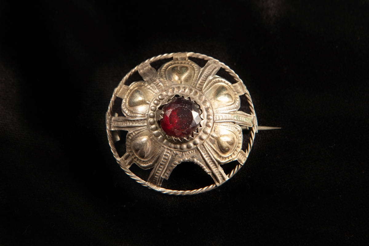 En rund malja av ciselerat och delvis genombrutet silver med dekor av hjärtan m.m. Infattad större sten av rött glas. Spännet, maljan, har gjorts om till en brosch.