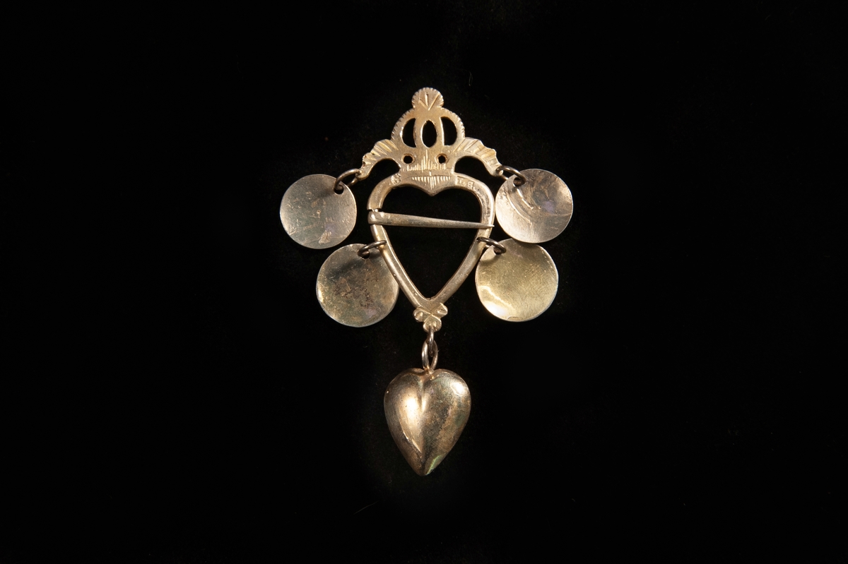 Hjärtformat spänne av förgyllt silver. Genombruten stiliserad krona upptill. Fyra skålar (varav de övre är graverade på ena sidan) hänger från spännet. Längst ner ett hjärta av silver (med annan stämpel: "E.E.., (Växjö) " (= Eric Ekström, Växjö, verksam cirka 1819-1840).