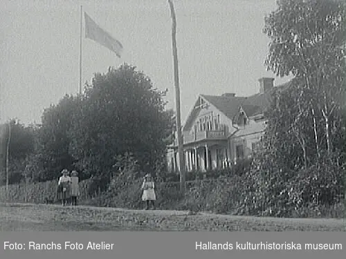 Fastigheten kallas Korsnäs och vägen är gamla E:6. Villan låg mitt emot Gästgiveriet i Slöinge. Exteriör. I förgrunden ses en kvinna med två barn.
