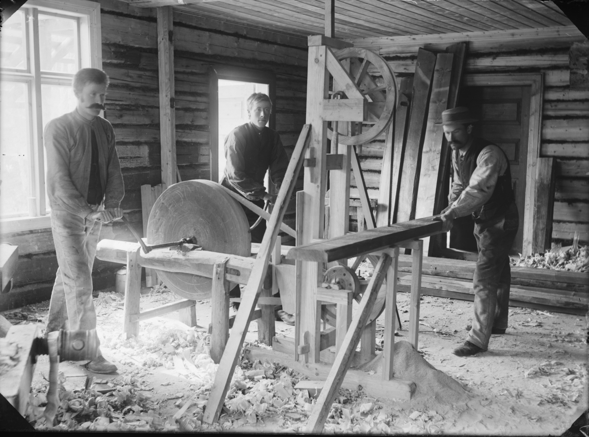 Snekkerverksted med hånddrevet bandsag. Interiør som viser to menn som drar en slipestein som igjen drar ei bandsag hvor en mann deler en planke.